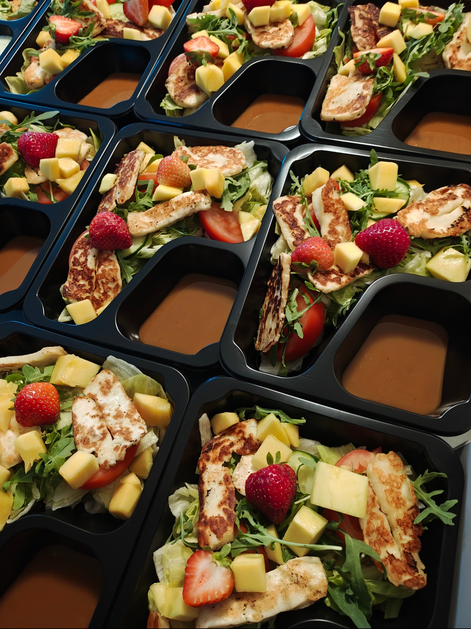 Sałatki z grillowanym serem halloumi, mango, truskawkami i sosem w czarnych pojemnikach cateringowych, ułożone równolegle. Zdrowe, kolorowe dania gotowe do spożycia.