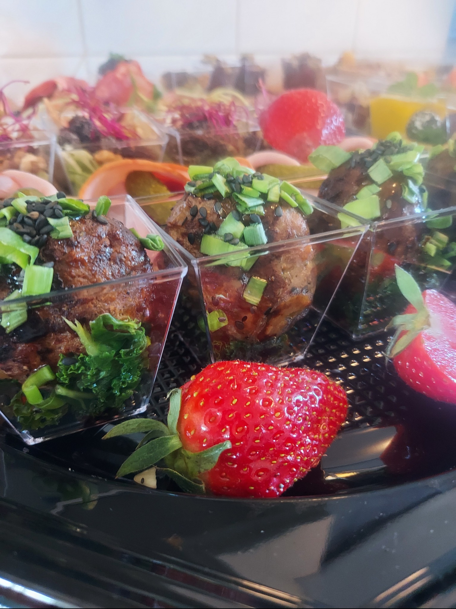 Elegancki catering: mini porcje mięsnych kulek z zielonymi dodatkami w przezroczystych naczyniach, uzupełnione świeżymi truskawkami na czarnej tacy.
