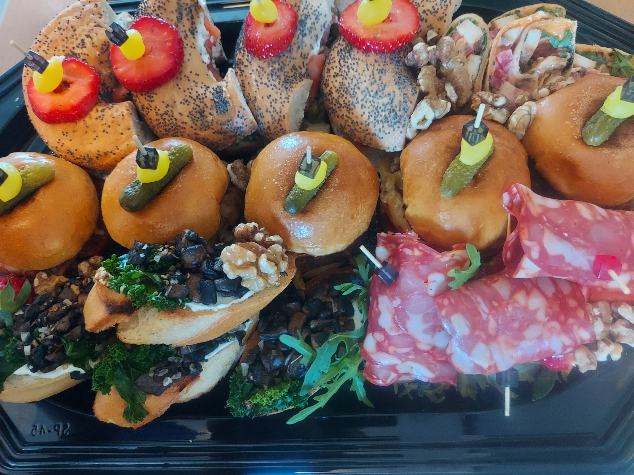 Elegancka taca cateringowa z mini burgerami, kanapeczkami z grzybami i tortillą, udekorowana owocami i warzywami, idealna na przyjęcia firmowe lub świąteczne.