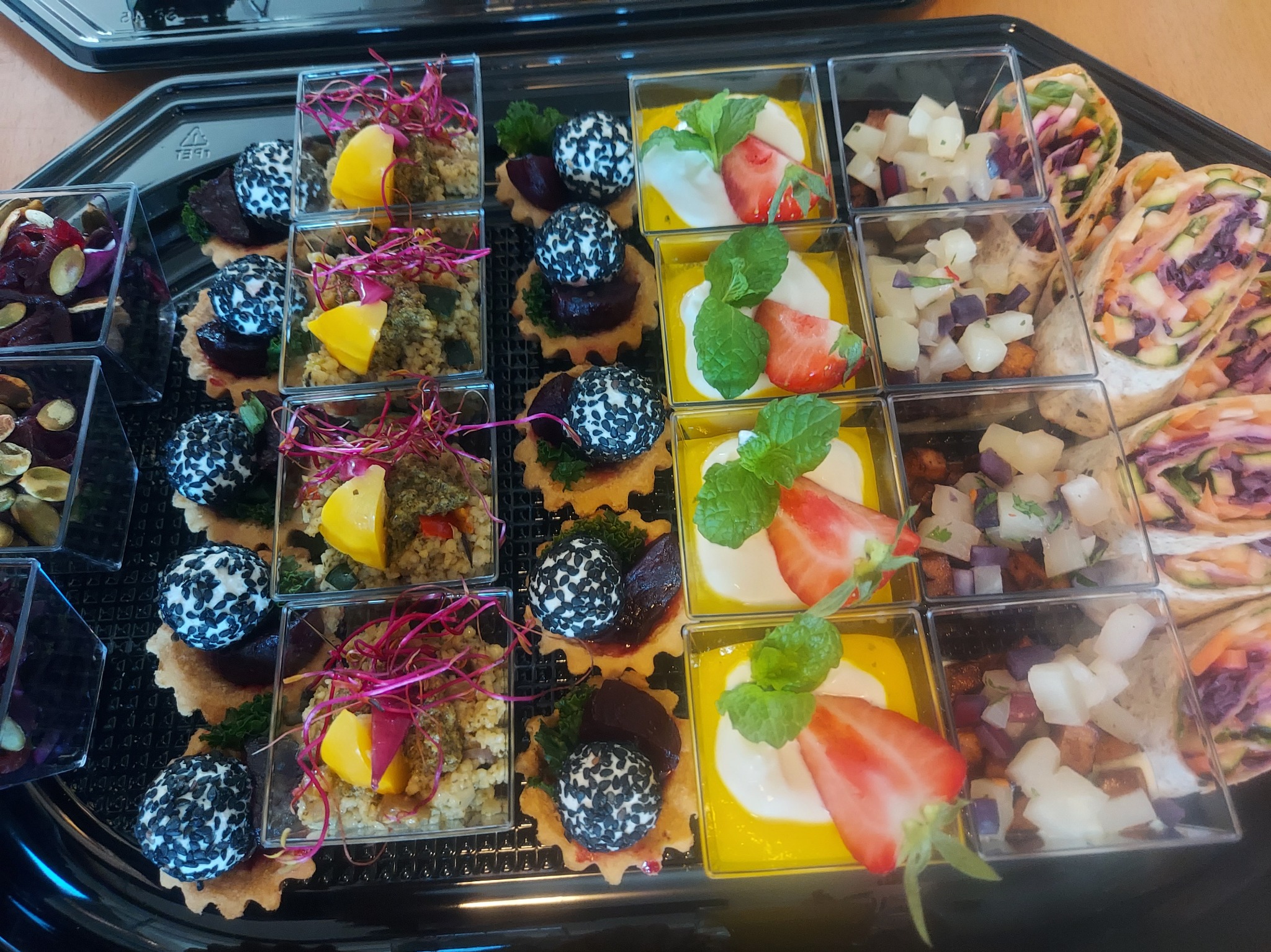 Elegancki catering: różnorodne przekąski w przezroczystych pojemnikach na czarnej tacy. Tartaletki, roladki i desery z owocami, warzywami i ziołami.