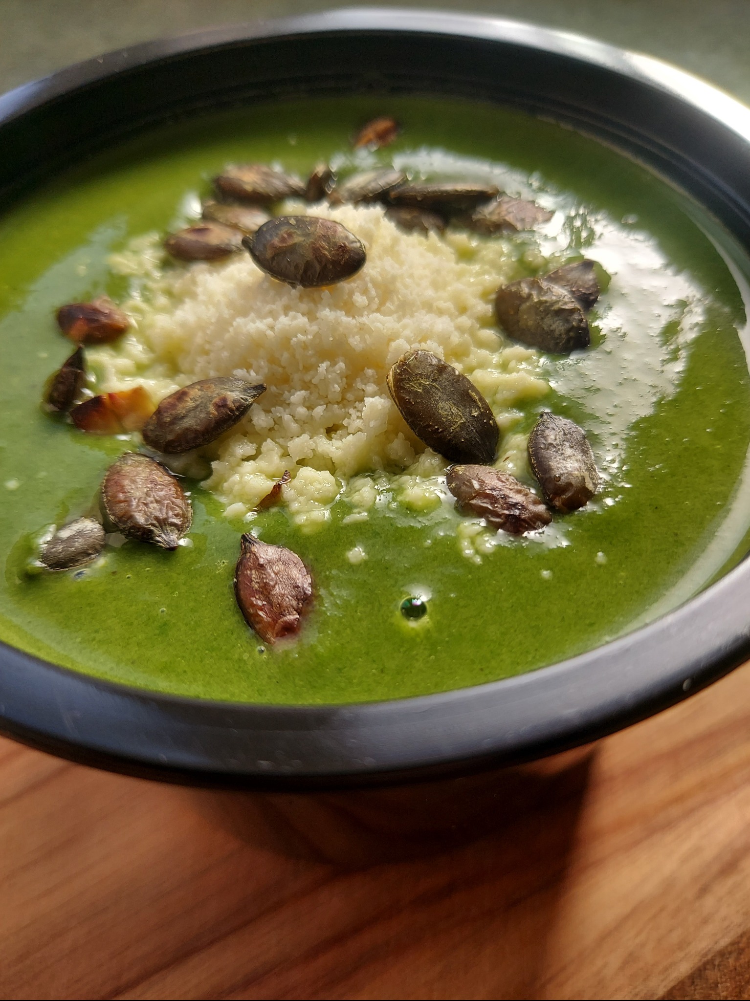 Zielony krem z zupy, posypany serem i pestkami dyni, w czarnej misce na drewnianym stole. Bliski kadr, ukazujący teksturę i składniki dania.