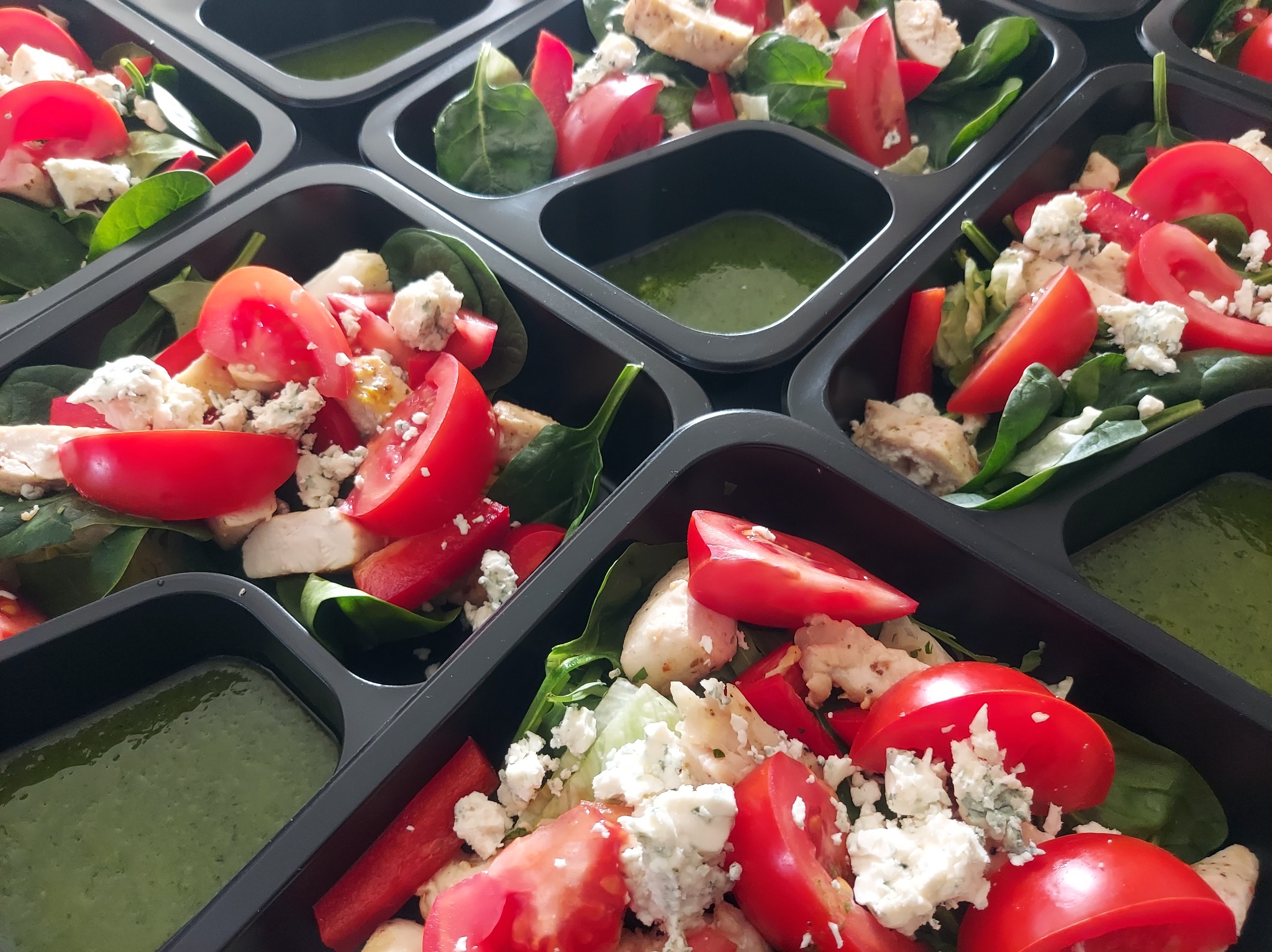 Sałatki z kurczakiem, pomidorami i serem pleśniowym w czarnych pojemnikach z zielonym sosem, gotowe do transportu. Catering dietetyczny.