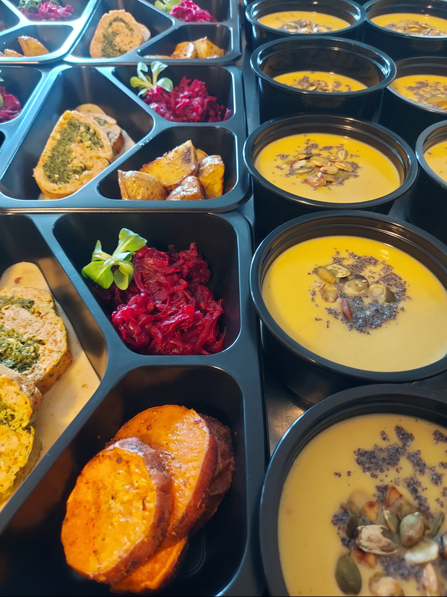 Zestaw cateringowy w czarnych pojemnikach: krem z dyni z pestkami, roladki szpinakowe, pieczone ziemniaki i buraczki. Gotowe dania na wynos.