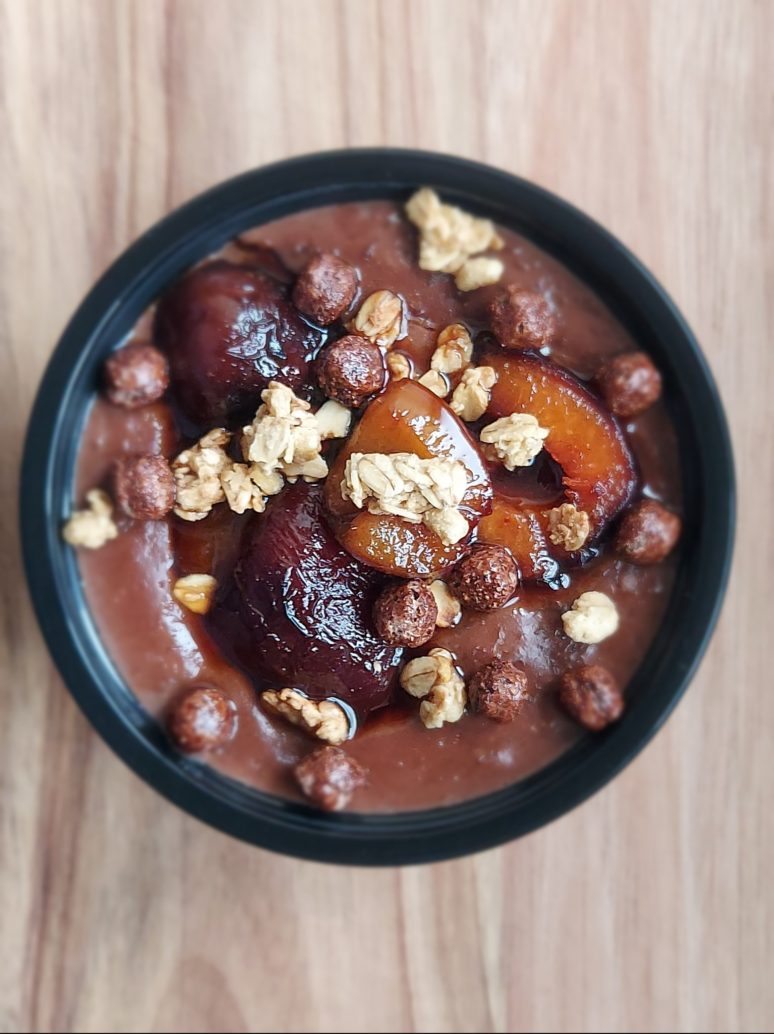 Deser w czarnej miseczce: brązowy krem, śliwki w syropie, płatki zbożowe i kulki czekoladowe. Widok z góry na drewnianym stole.