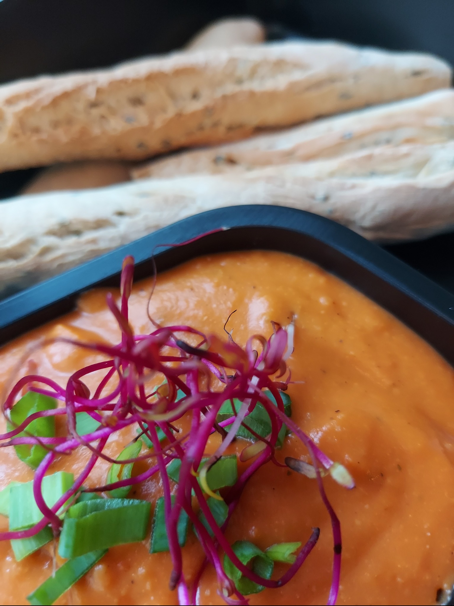 Krem z warzyw w czarnym naczyniu, udekorowany zielonym szczypiorkiem i różowymi kiełkami. W tle bagietki.