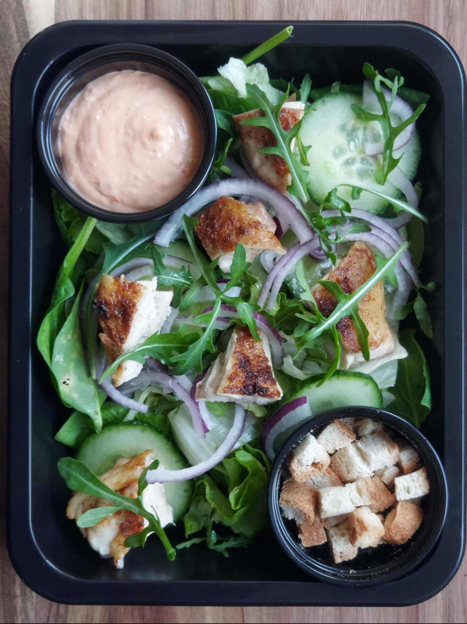 Sałatka z grillowanym kurczakiem, ogórkiem, czerwoną cebulą i grzankami, serwowana w czarnym pojemniku z sosem w oddzielnej miseczce.