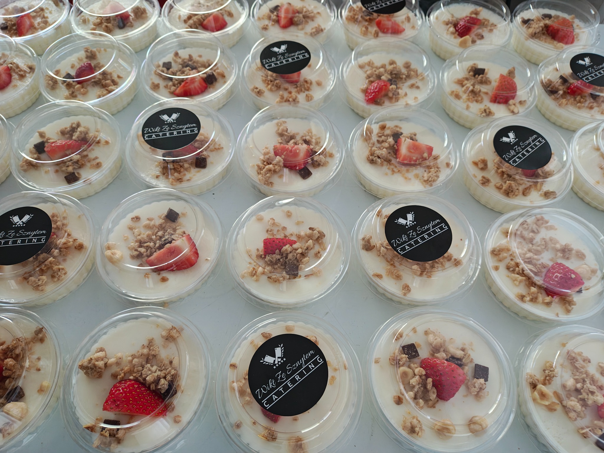Deser w plastikowych pojemnikach z owocami i granolą, ułożony w rzędach. Widoczne logo firmy cateringowej na niektórych pojemnikach. Słodki poczęstunek.