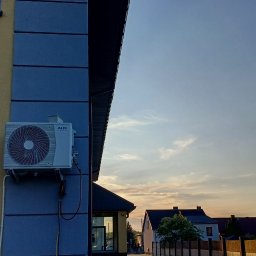 Agadi Przemysław Blukacz - Zewnętrzna jednostka klimatyzacji AUX zamontowana na elewacji budynku z widocznymi przewodami i manometrami serwisowymi; w tle zabudowa jednorodzinna i niebo o zmierzchu.