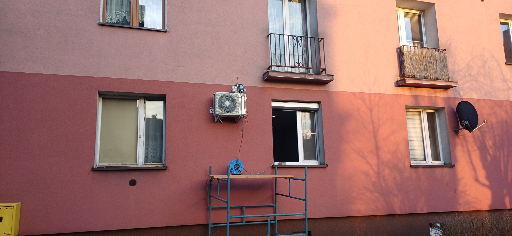 Zewnętrzna jednostka klimatyzacji zamontowana na ścianie dwukondygnacyjnego budynku mieszkalnego, podłączona do przewodów serwisowych i niebieskiego zwijadła kabla na rusztowaniu.