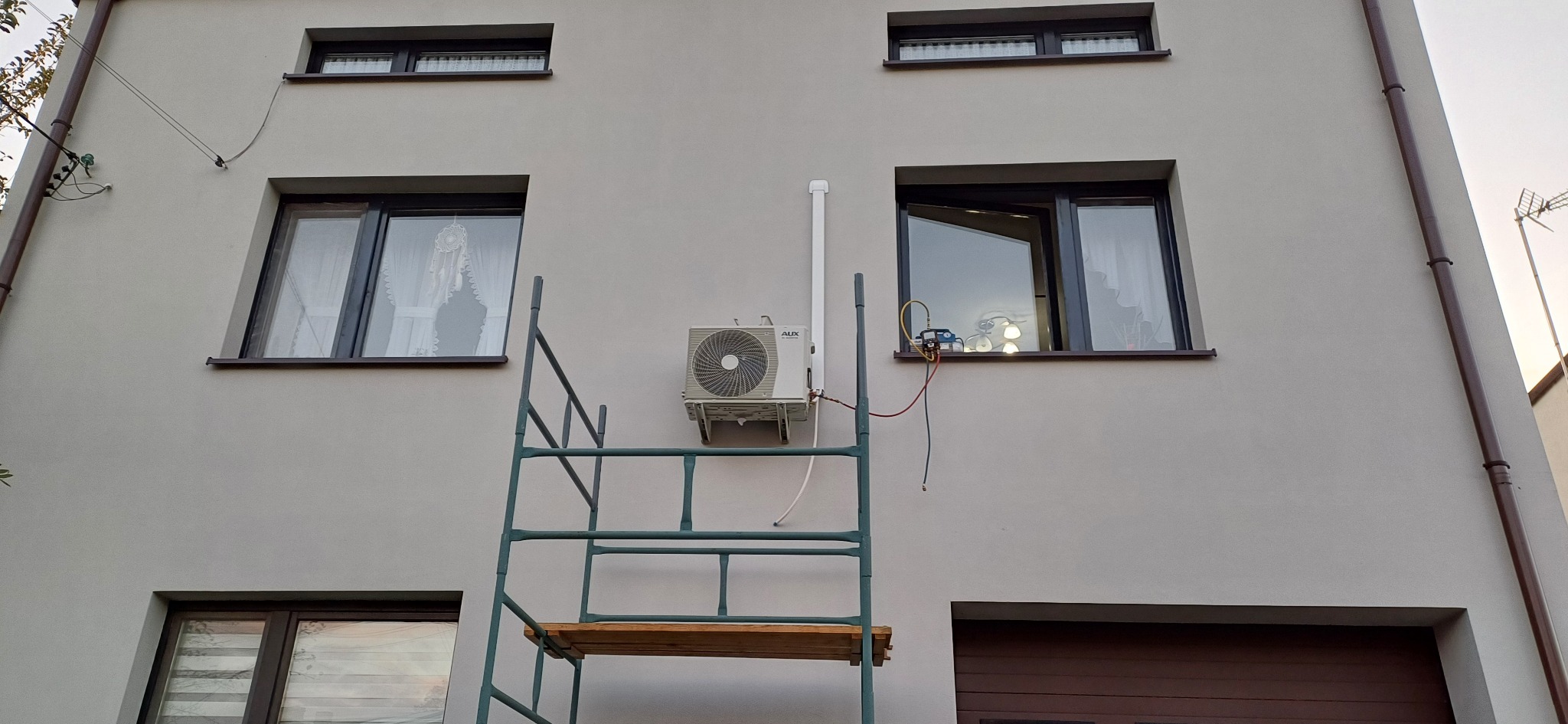Zewnętrzna jednostka klimatyzacji AUX zamontowana na ścianie domu, widoczna z rusztowania, podłączona do manometrów serwisowych, okno z otwartą ramą w tle.