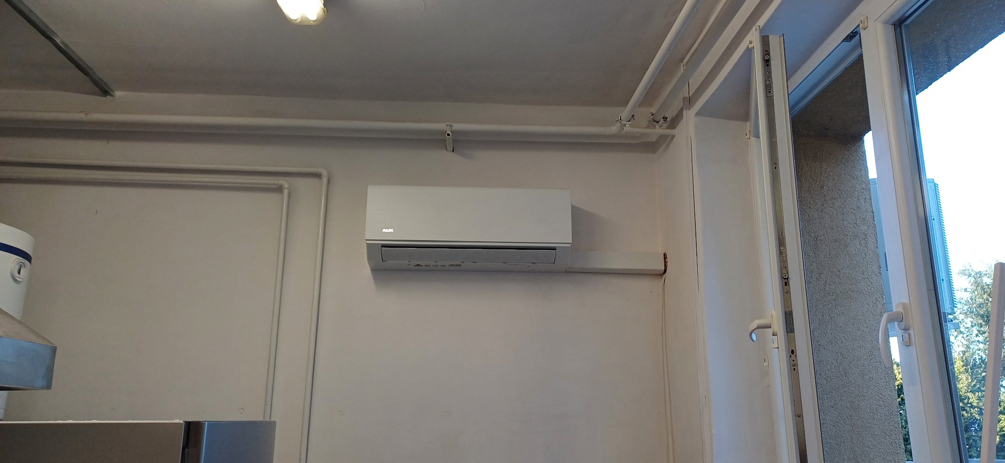 Biała jednostka klimatyzacyjna marki AUX zamontowana na ścianie w pomieszczeniu, z widocznymi rurami instalacyjnymi pod sufitem i fragmentem otwartego okna balkonowego.