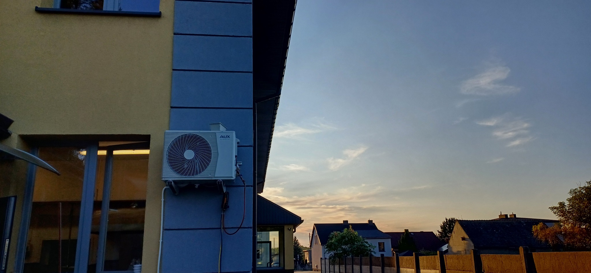 Zewnętrzna jednostka klimatyzacji AUX zamontowana na elewacji budynku z widocznymi przewodami i manometrami serwisowymi; w tle zabudowa jednorodzinna i niebo o zmierzchu.