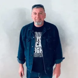 Portret uśmiechniętego mężczyzny z siwą brodą, w dżinsowej kurtce i czarnym t-shircie z napisem, stojącego na tle jasnej ściany.