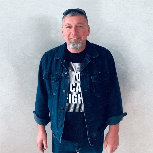 Portret uśmiechniętego mężczyzny z siwą brodą, w dżinsowej kurtce i czarnym t-shircie z napisem, stojącego na tle jasnej ściany.