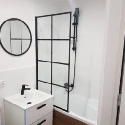 Nowoczesna łazienka z białą umywalką na szafce z czarnymi uchwytami, okrągłym lustrem w czarnej ramie, kabiną prysznicową z czarnymi profilami i brązowymi płytkami podłogowymi.