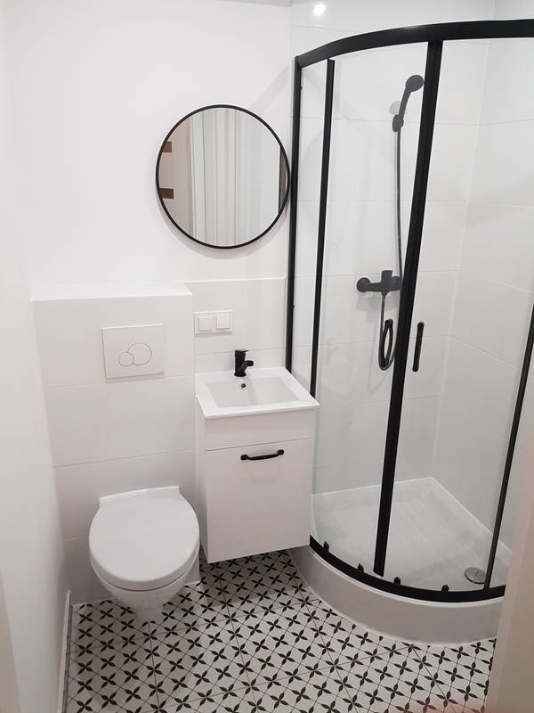 Nowoczesna łazienka w bieli i czerni z kabiną prysznicową o czarnych profilach, okrągłym lustrem i wzorzystą podłogą.