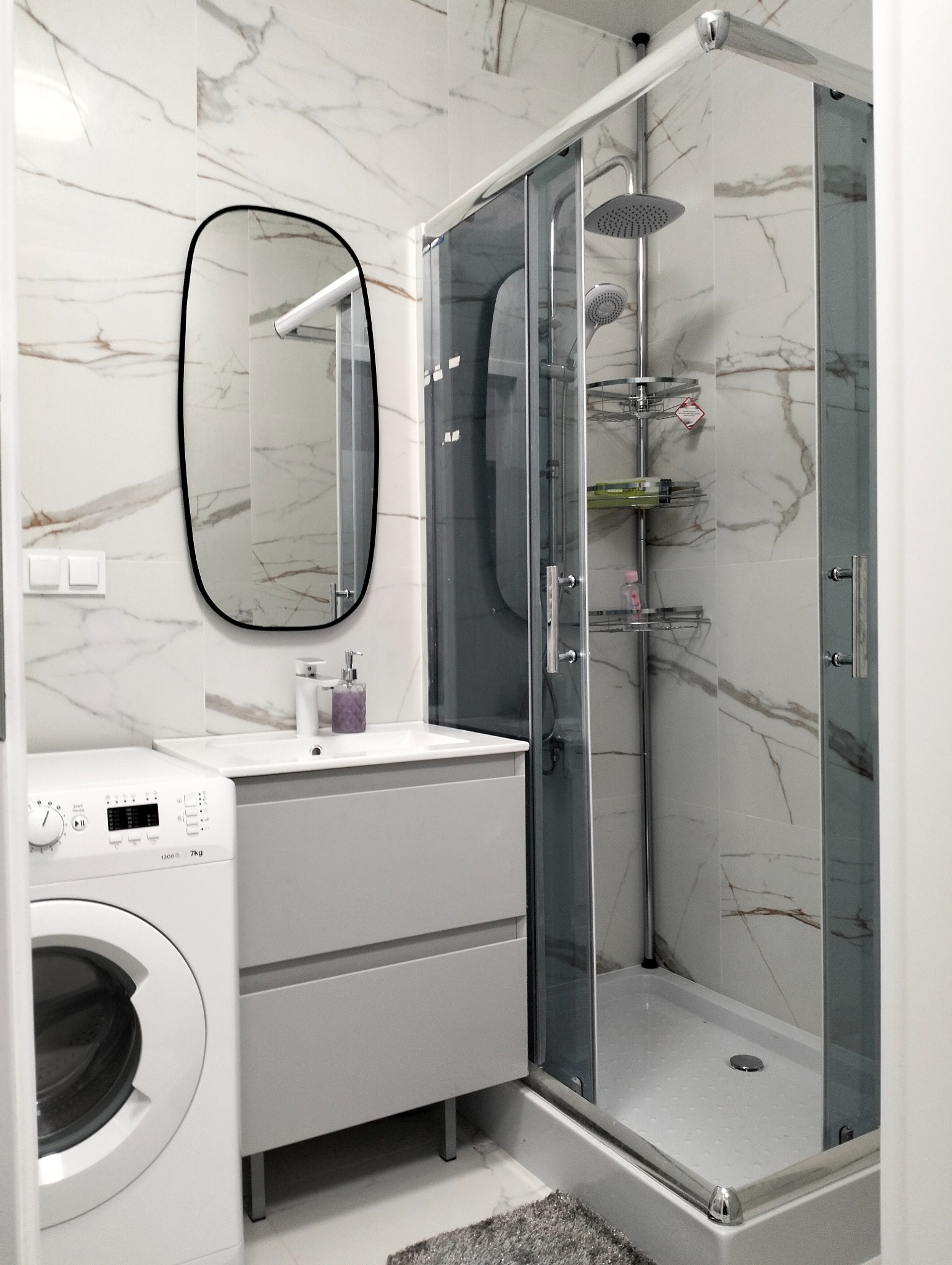 Nowoczesna łazienka z kabiną prysznicową, umywalką z szafką w odcieniach szarości, pralką i lustrem o zaokrąglonym kształcie na tle marmurowych płytek.