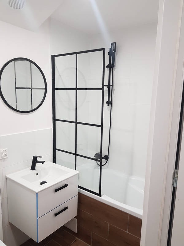 Nowoczesna łazienka z białą umywalką na szafce z czarnymi uchwytami, okrągłym lustrem w czarnej ramie, kabiną prysznicową z czarnymi profilami i brązowymi płytkami podłogowymi.