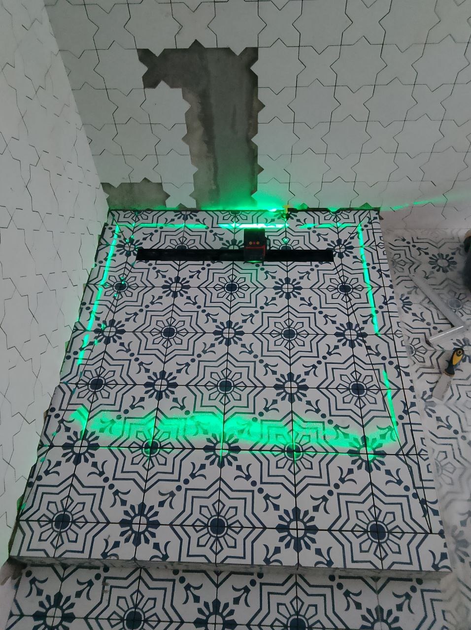 Świeżo ułożone, wzorzyste płytki w kabinie prysznicowej, podświetlone zielonym laserem, z widocznym odpływem liniowym i niedokończonym sufitem.