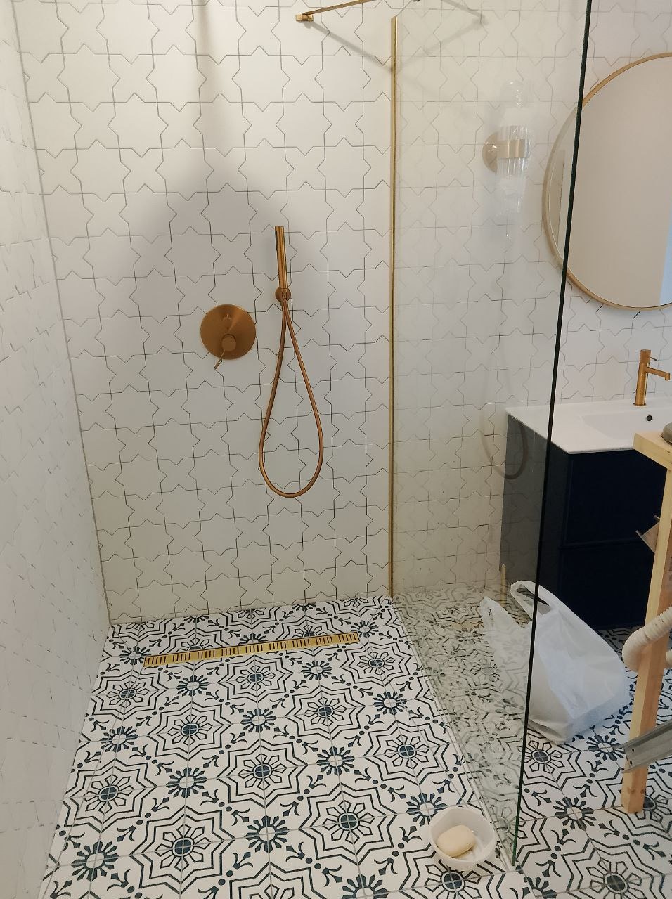 Wykończona łazienka z kabiną prysznicową typu walk-in, biało-czarną wzorzystą podłogą, złotą armaturą i odpływem liniowym. Na ścianach białe płytki z delikatnym szarym wzorem.