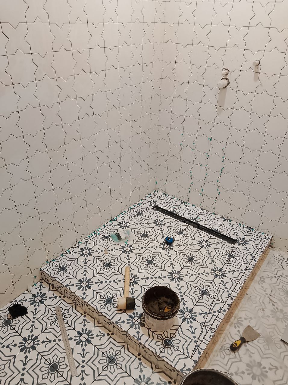 Wnętrze łazienki w trakcie prac wykończeniowych: ułożone płytki podłogowe z wzorem kwiatowym, widoczny odpływ liniowy, ściany przygotowane pod okładzinę z płytek o geometrycznym wzorze, narzędzia...