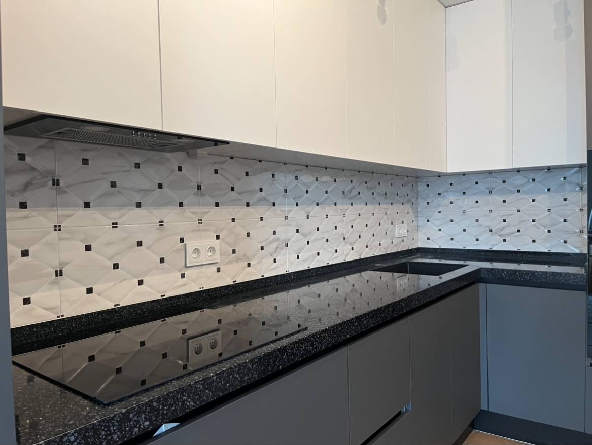 Wykończona kuchnia z białymi szafkami górnymi i szarymi dolnymi, czarny blat z wbudowaną płytą i wzorzyste płytki na ścianie pomiędzy szafkami.