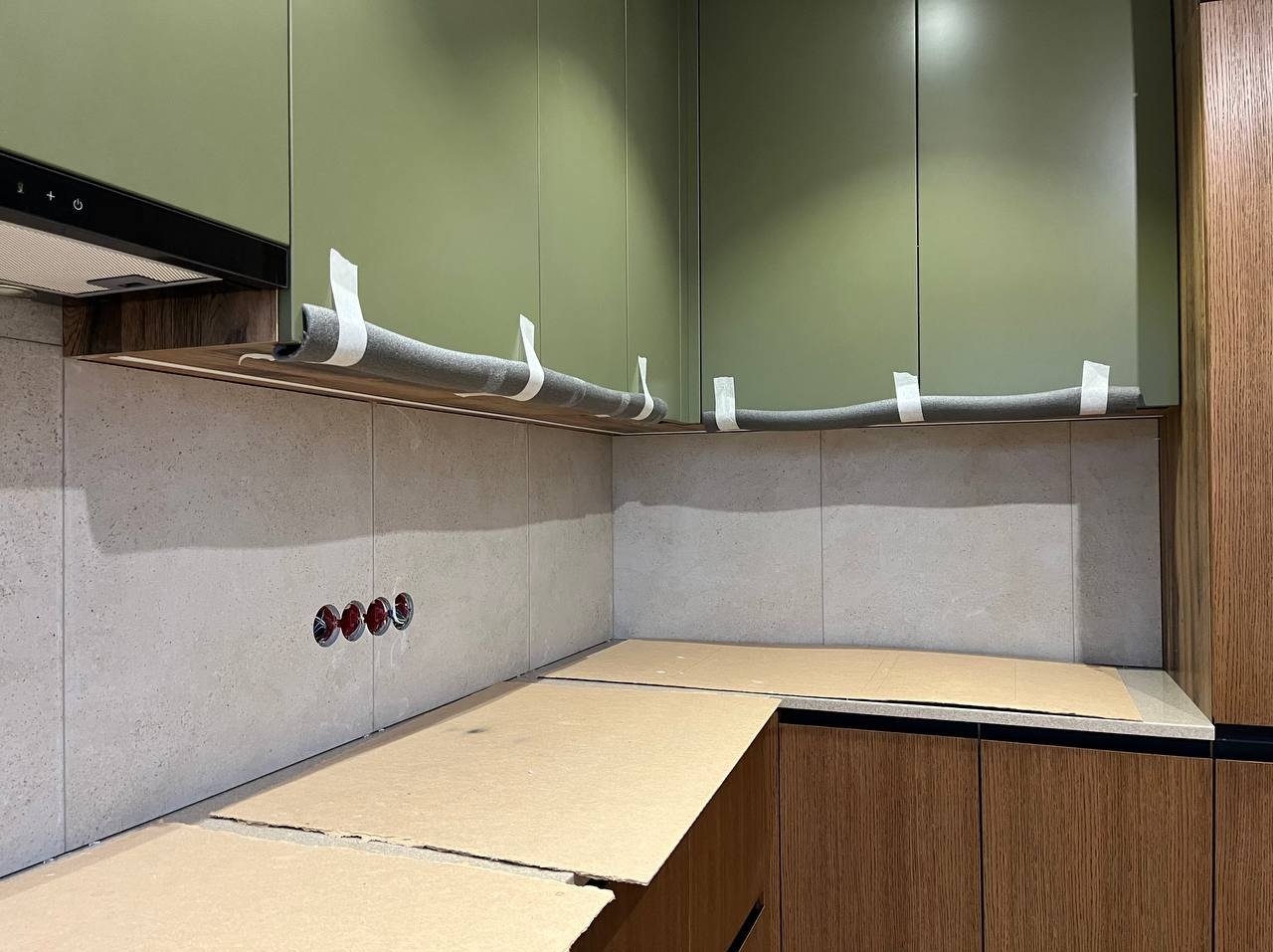 Kuchnia w trakcie wykańczania: oliwkowe szafki wiszące zabezpieczone pianką, jasne płytki na ścianie z widocznymi gniazdkami, blaty zakryte kartonem, dolne szafki z drewnopodobnym fornirem.