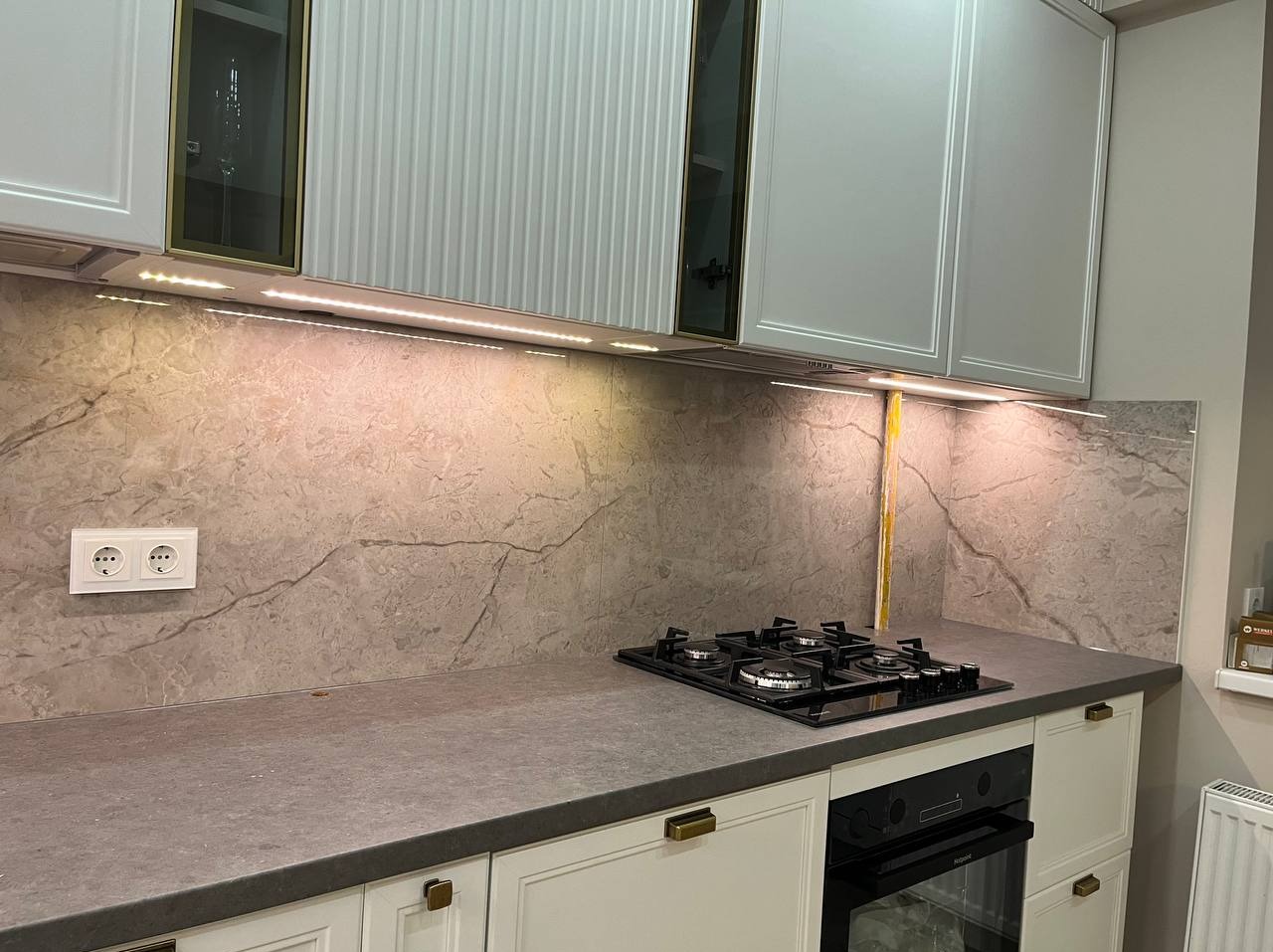 Wykończona kuchnia z szarym blatem, marmurową ścianą i białymi szafkami z złotymi uchwytami, oświetlona taśmą LED pod szafkami wiszącymi.