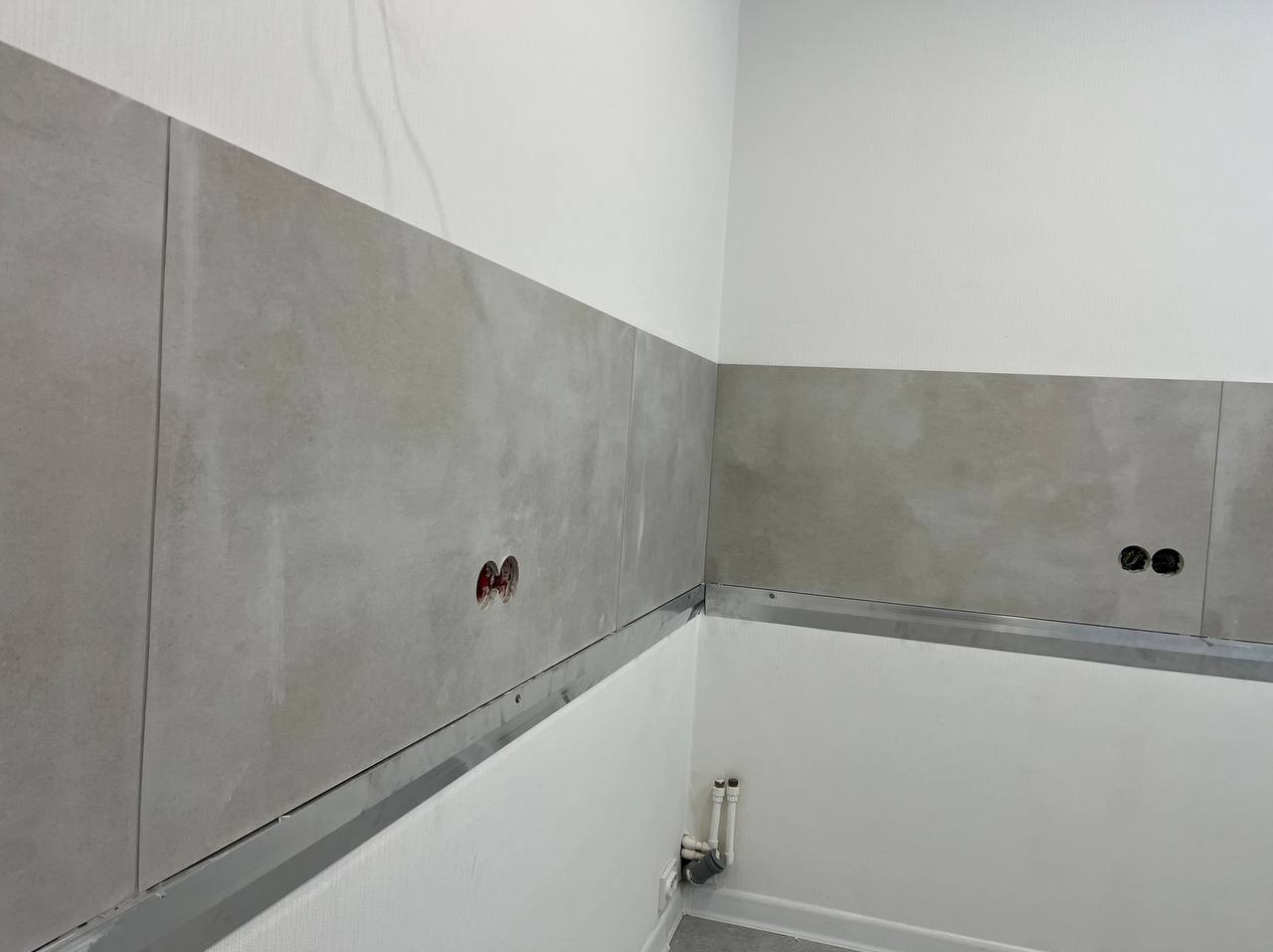 Ściana w trakcie wykańczania z szarymi płytkami, widoczne otwory na gniazdka elektryczne, metalowa listwa zabezpieczająca, białe ściany i rury kanalizacyjne przy podłodze.