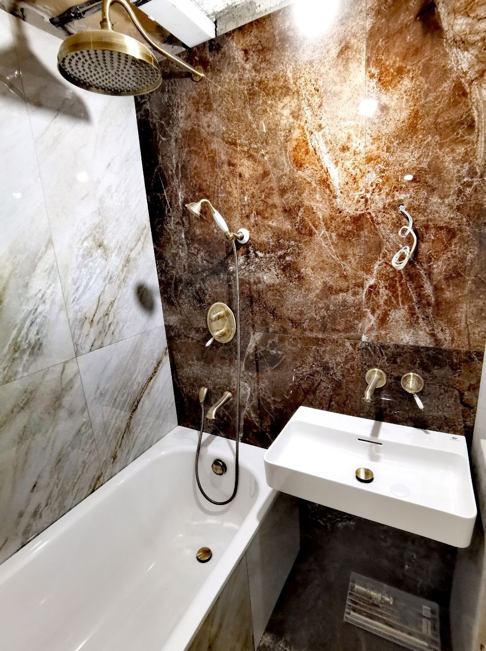 Nowoczesna łazienka z marmurowymi płytkami w odcieniach brązu i bieli, wyposażona w białą wannę, umywalkę o minimalistycznym designie i armaturę w kolorze starego złota.