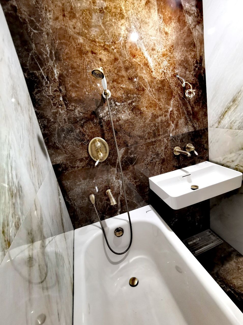 Biała wanna z armaturą w kolorze starego złota na tle ściany z brązowego marmuru, minimalistyczna umywalka nablatowa.