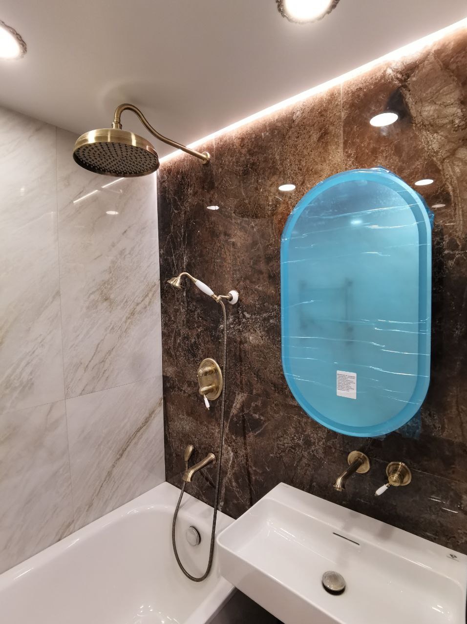 Nowoczesna łazienka z elementami w stylu retro: złota armatura, marmurowe płytki na ścianach, umywalka z prostokątną misą, lustro zabezpieczone folią ochronną.