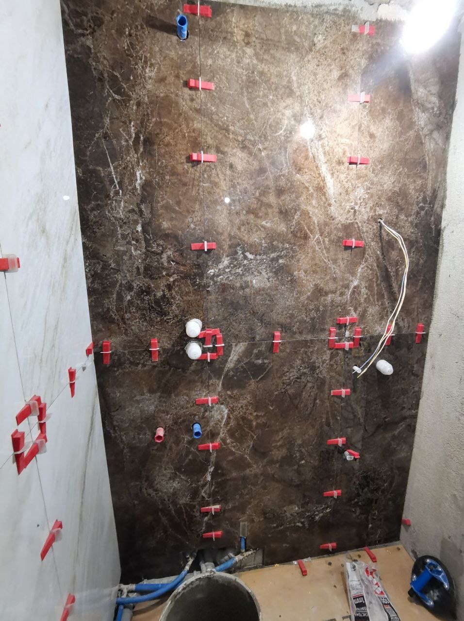 Montaż brązowych płytek imitujących marmur na ścianie z użyciem czerwonych krzyżyków dystansowych i poziomicy laserowej; widoczne rury instalacji wodnej i kanalizacyjnej.