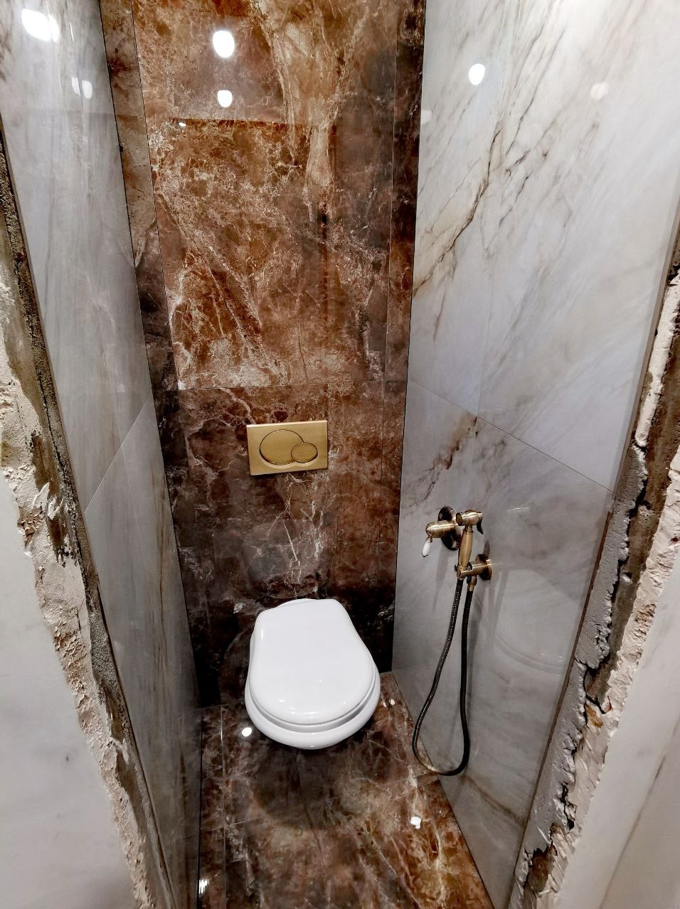 Niewielka toaleta w trakcie remontu, widoczne marmurowe płytki w odcieniach brązu i szarości, podwieszana miska WC, złota armatura i częściowo wykończone ściany.