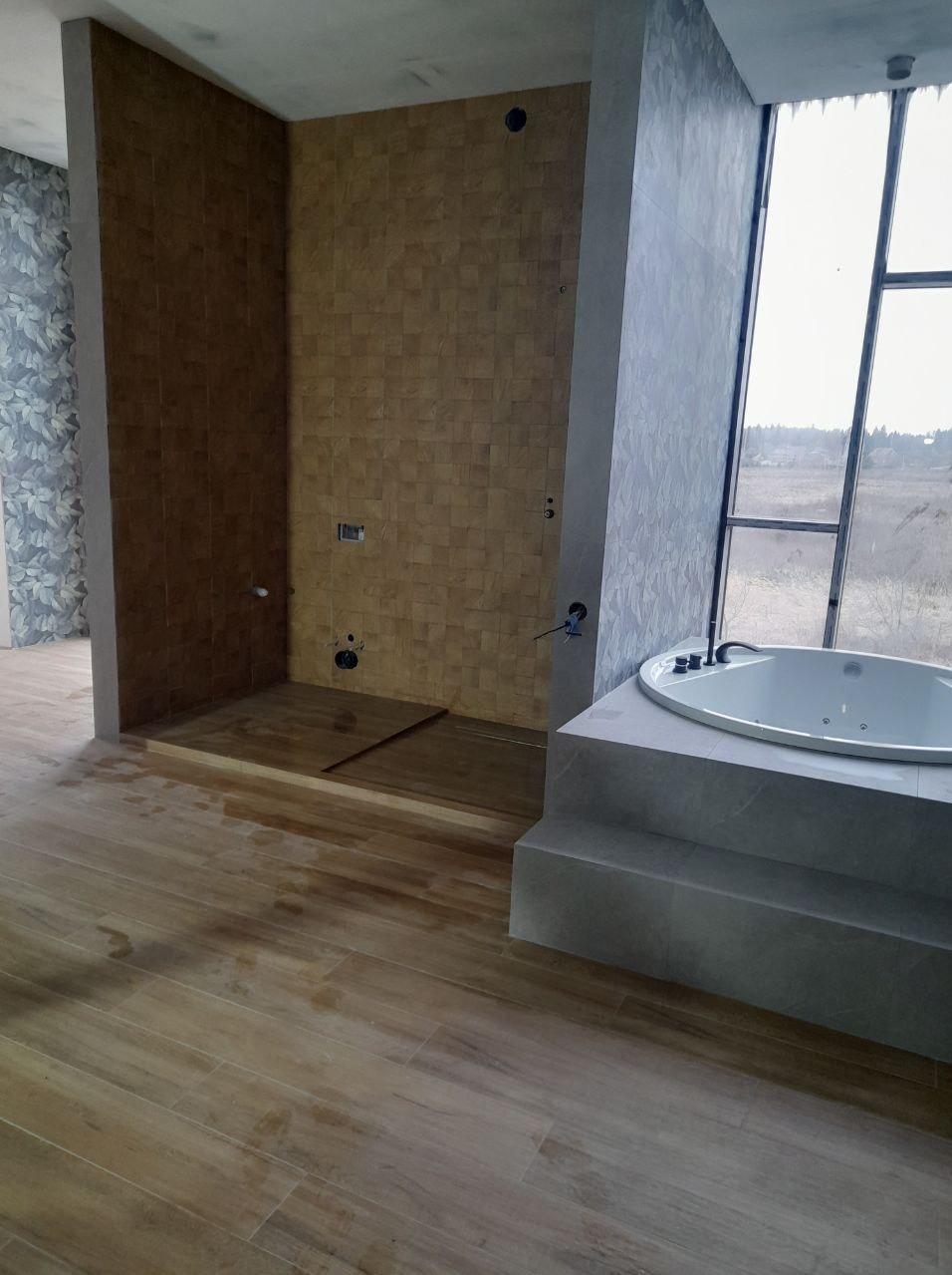 Wnętrze łazienki w trakcie wykończenia z widoczną kabiną prysznicową wyłożoną mozaiką drewnopodobną, wolnostojącą wanną przy oknie i drewnopodobną podłogą.