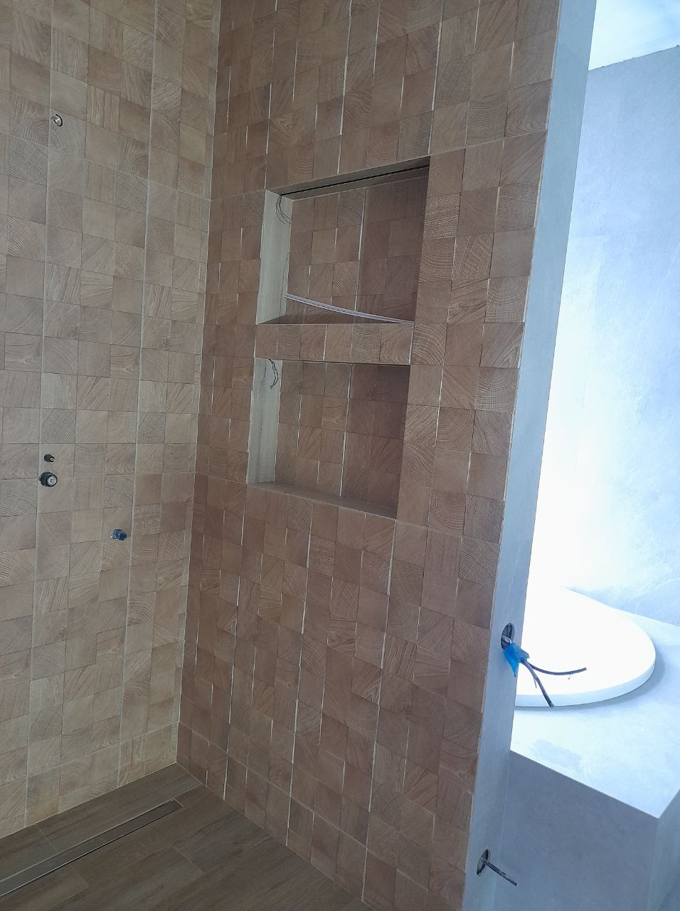 Ściana w łazience wykończona drewnopodobnymi, kwadratowymi płytkami z wnękami, widoczne przyłącza do armatury i odpływ liniowy w podłodze.