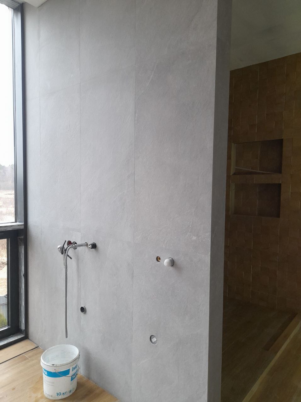 Ściana łazienki wyłożona szarymi płytkami wielkoformatowymi, widoczny fragment instalacji prysznicowej i przygotowane punkty podłączeń, wiaderko z wodą i gąbką przy ścianie, w głębi kabina...