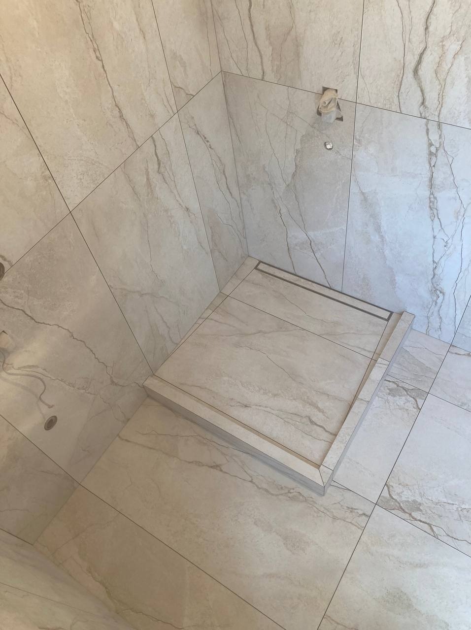 Wykończony brodzik w kabinie prysznicowej z jasnymi, marmurkowymi płytkami, widoczny odpływ liniowy i przygotowane punkty instalacyjne na ścianie.