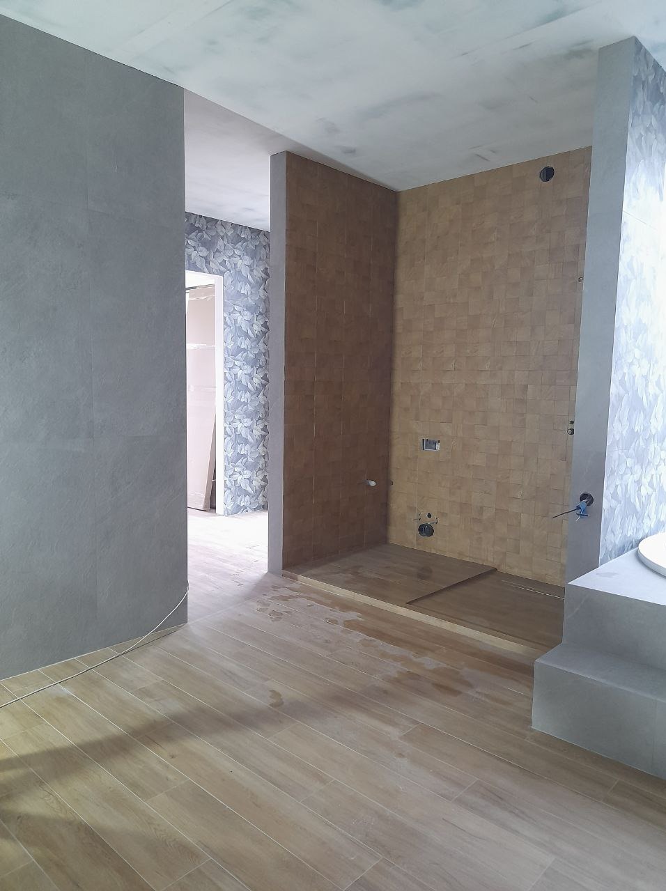 Wnętrze łazienki w trakcie remontu, widoczne beżowe płytki na ścianach kabiny prysznicowej, drewnopodobna podłoga, szare ściany i fragment ściany z tapetą w kwiaty w tle, surowy stan wykończenia.