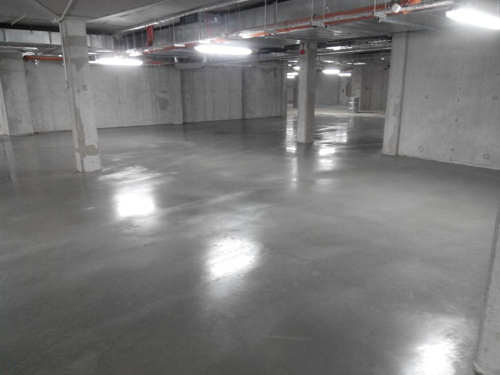 Gładka, szara posadzka w surowym, betonowym wnętrzu hali przemysłowej, oświetlona jarzeniówkami, z widocznymi słupami nośnymi i instalacjami pod sufitem.