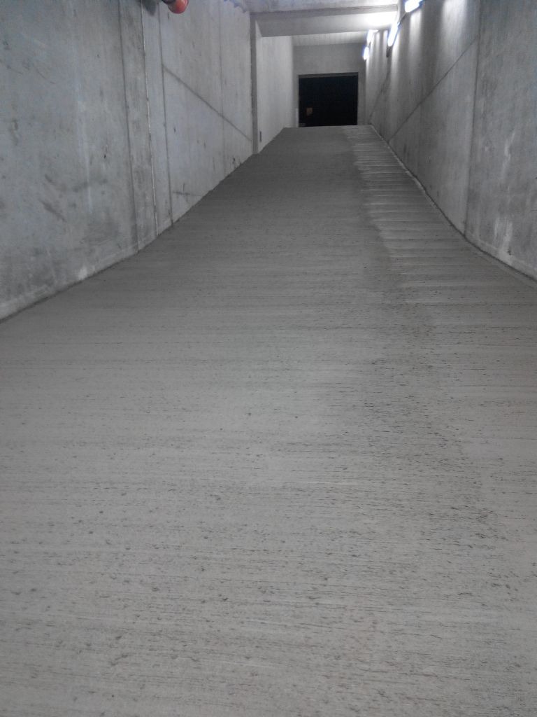 Betonowa rampa wewnątrz budynku z szarymi ścianami i oświetleniem LED u góry, prowadząca do ciemnego wejścia.