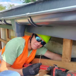 Monter w kasku i kamizelce, z narzędziami, instaluje rynny na dachu budynku. Widoczny fragment dachu, rynny, drewniana konstrukcja i narzędzia pracy.