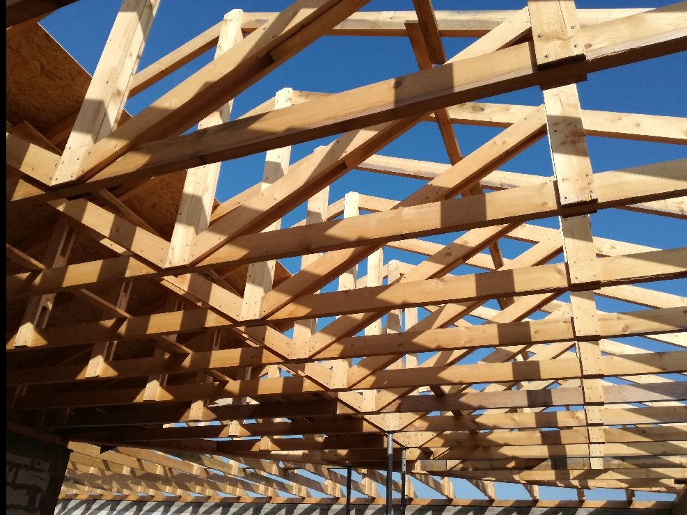 Konstrukcja drewniana dachu w trakcie budowy, widok od wewnątrz na niebieskie niebo. Elementy połączone, widoczne cienie i geometria belek.
