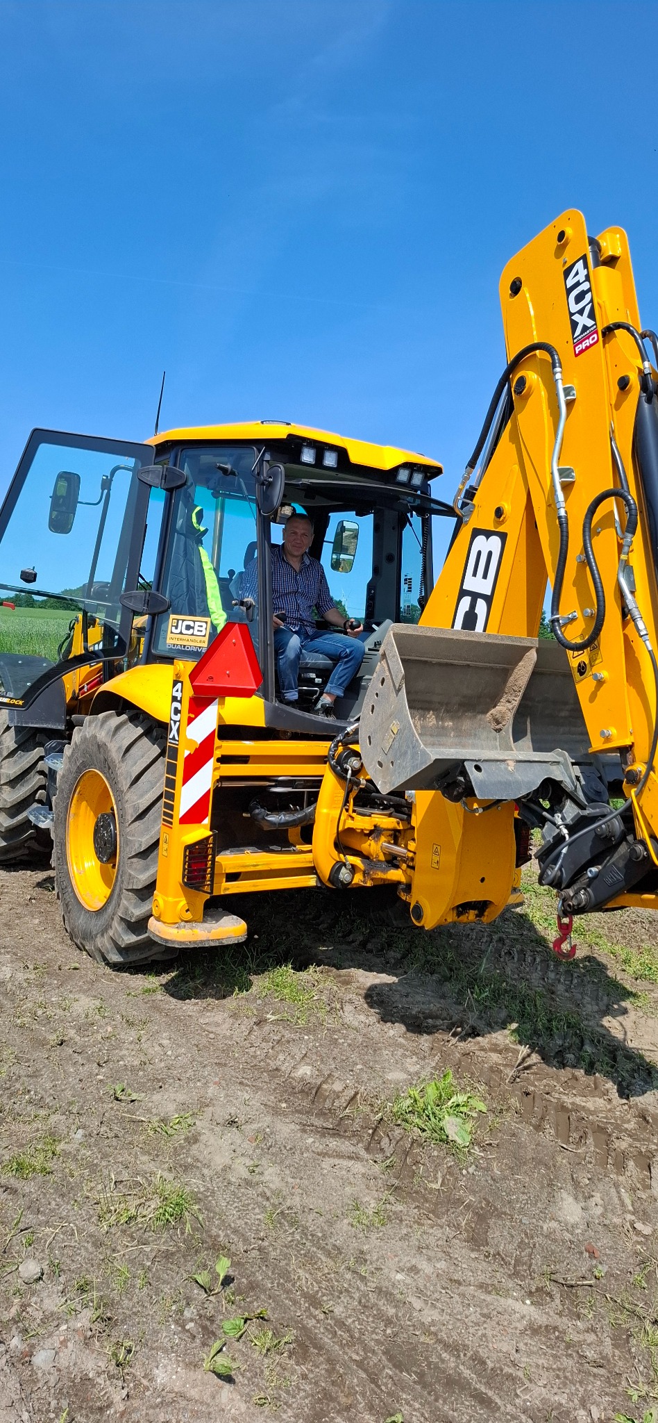 Operator w kabinie żółtej koparko-ładowarki JCB 4CX, na tle błękitnego nieba. Widoczne ramię koparki i opony. Praca w terenie.