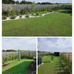 Zadbany ogród z równo przystrzyżonym trawnikiem, rabatami z białym kamieniem i różnorodną roślinnością, ogrodzony siatką z widokiem na okolicę. Słoneczny dzień.