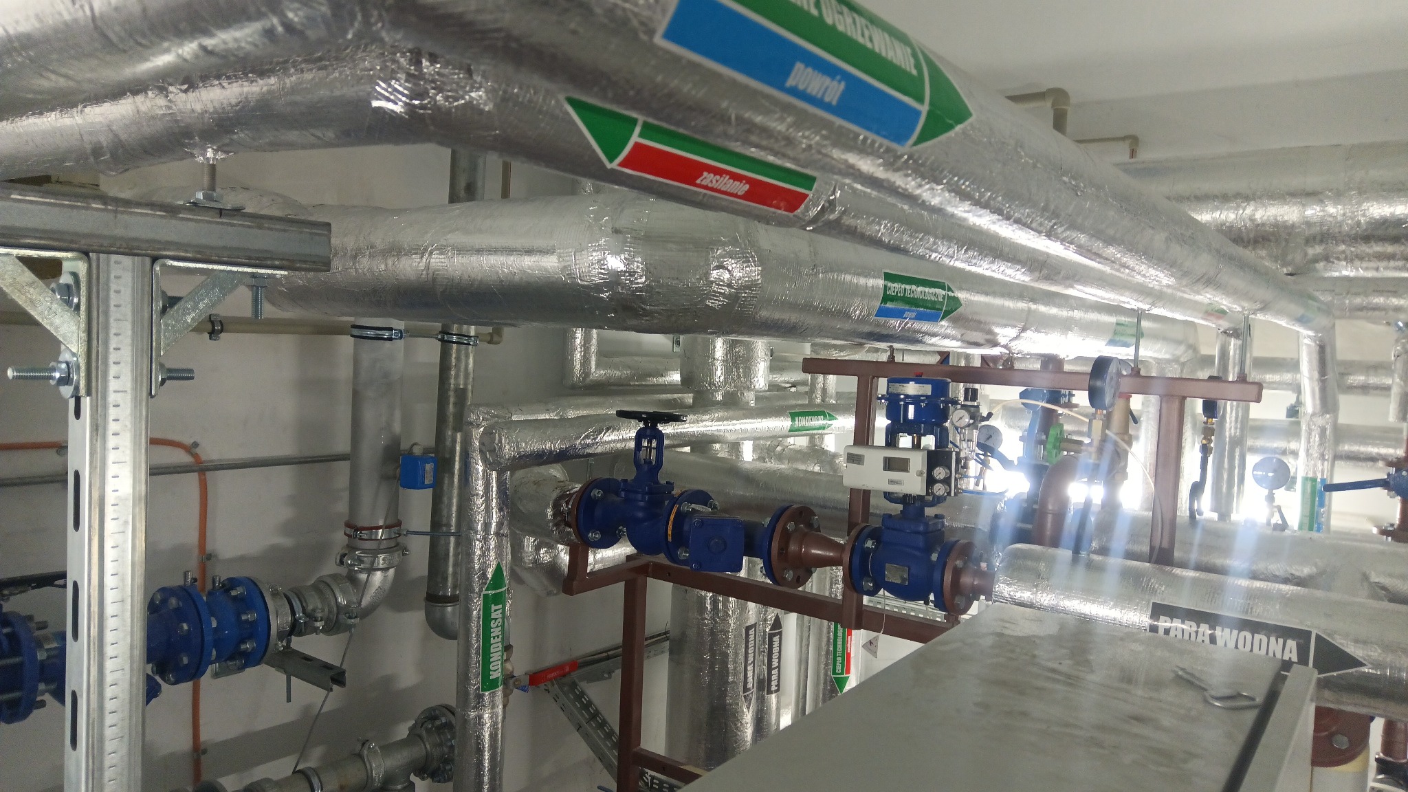 Złożona instalacja rur z izolacją termiczną, zaworami i oznaczeniami (np. 'Para wodna', 'Zasilanie'). Widoczne elementy montażowe i wsporniki. Ujęcie z dołu, oświetlenie techniczne.