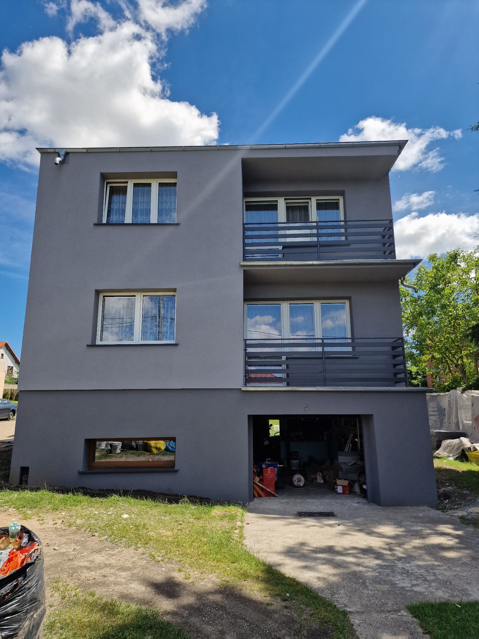 Dwupiętrowy dom z szarą elewacją, balkonami z metalowymi barierkami i widocznym garażem, uchwycony w słoneczny dzień z lekkim bałaganem na trawniku.