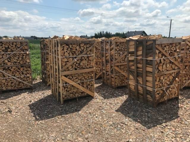 Ułożone w drewnianych skrzyniach pocięte kawałki drewna na zewnątrz, na żwirowym podłożu, w tle widoczne budynki i słup energetyczny.