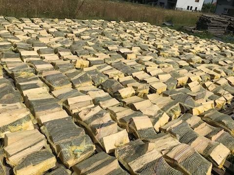 Szeroki kadr na ułożone w rzędach worki z połupanym drewnem na paletach, suszące się na zewnątrz pod słońcem, z trawą i budynkiem w tle.