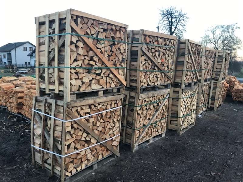 Ułożone w drewnianych skrzyniach, pocięte kawałki drewna na paletach, zabezpieczone sznurkiem, składowane na zewnątrz, w tle budynek mieszkalny i drzewo.